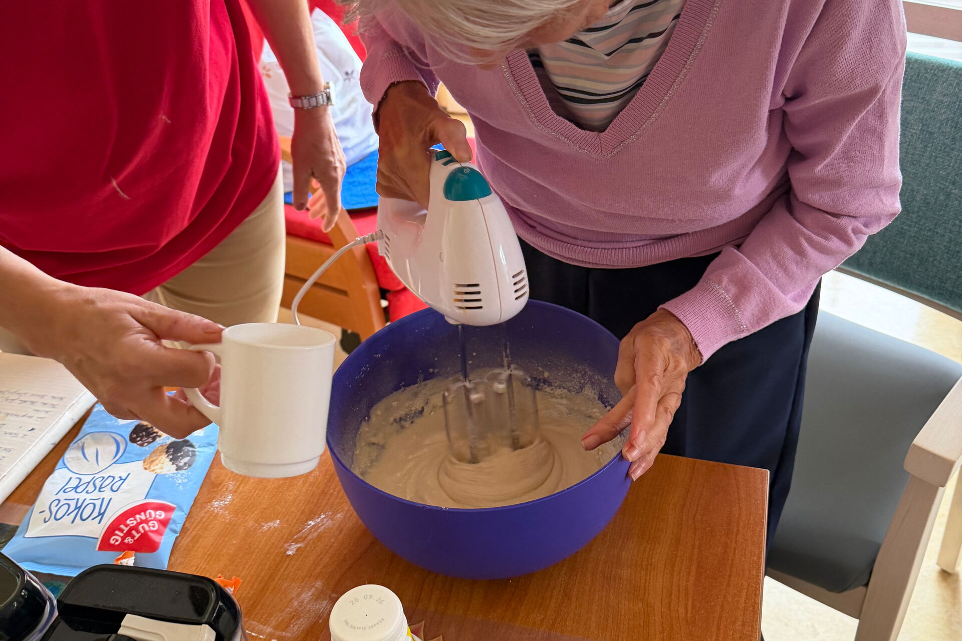 Seniorin rührt mit Rührgerät Teig in einer blauen Schüssel. Pflegerin steht neben ihr.