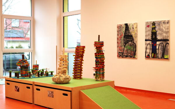 Lichtdurchfluteter Raum. Zwei Bilder hängen nebeneinander an der Wand. Im Zentrum Türme aus Holz und anderem Material.