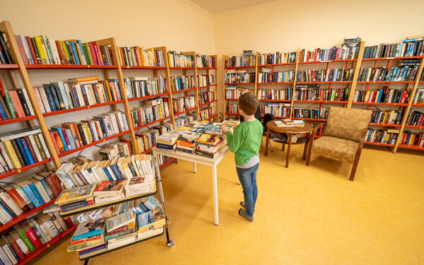 Kind steht an einem Tisch und schaut sich ein Buch an. Bibliothek, Regale mit vielen Büchern stehen an den Wänden.