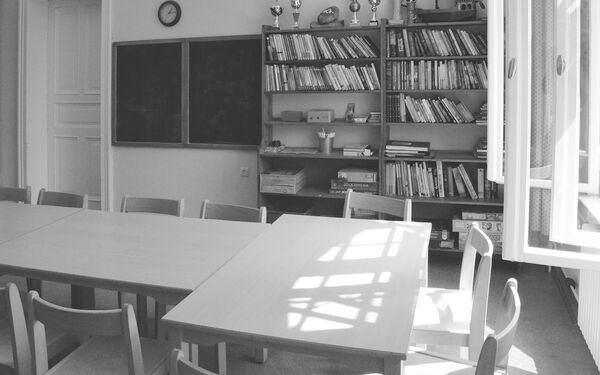 Raum mit geöffneten Fenstern. Tische und Stühle. An der Wand ein großes Regal mit vielen Büchern und Spielen.