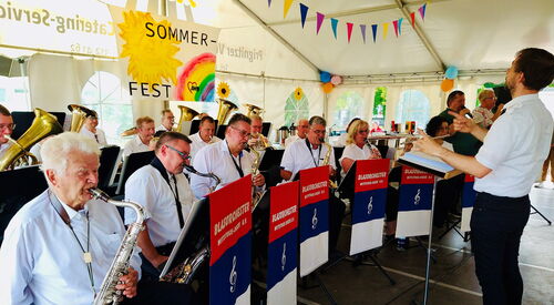 Musiker mit Blasinstrumenten und rechts der Dirigent, alle tragen weiße Hemden uns schwarze Hosen.