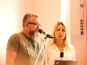 Finger vor der Linse. Ein Mann und eine Frau am Rednerpult. Mann mit Brille und Bart, spricht in Mikrophon.