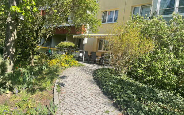 Barrierearmer Zugang über den Garten auf die Terrasse. Es stehen Stühle und Tische auf der Terrasse und eine Markise spendet Schatten.