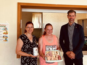 Gruppenfoto: 3 Personen, Frau im Sommerkleid mit Spendendose in der Hand, Frau mit hält Tablett mit belegten Broten, großer Mann im dunklen Anzug und grauen Hemd.
