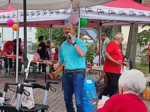 Rote AWO Pavillons, Helfer in roten AWO T-Shirts. Im Mittelunkt Mann mit Mikrofon. Der Mann har eine hohe Stirn, sonst kurze graue Haare, er trägt ein blaues kurzärmliges Hemd und braue Jeans.