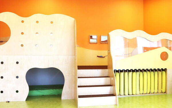 Heller Raum, orangene Wand, grüner Fußboden. Dominant eine heller Holz-Kletter-Versteck-Liege Konstruktion, wo unten Stauraum für Matratzen ist.