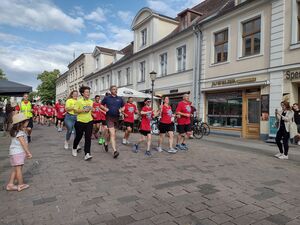 Eine große Gruppe Menschen in roten T-Shirts und schwarzen kurzen Hosen, läuft / rennt eine Straße hinauf. Daneben der Potsdamer Oberbürgermeister in Sportkleidung rennt mit.