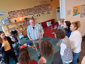 In einem Kita-Raum mit Spielzeug auf dem Fußboden. Die Gruppe Frauen steht im Halbkreis um einen Mann. Mann ist etwas älter, kurze Haare, Brille. Er trägt ein bunt längsgestreiftes helles Hemd.