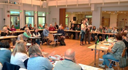 Sehr großer Raum, Tische zu einem großen Viereck gestellt an denen vielen Menschen sitzen. In deren Mitte stehen eine Frau und mit Mikro und ein Mann.