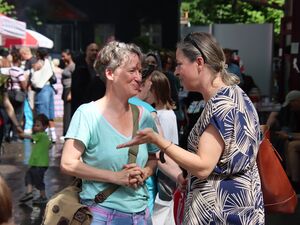 Draußen, Sonne scheint. Zwei Frauen unterhalten sich. Ringsherum viele Menschen und Trubel. Frauen tragen sommerliche Kleidung.