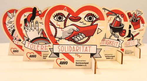 Tischaufsteller. In Herzform. Ganz vorn Solidarität, dahinter versetzt Toleranz und Gerechtigkeit. Dahinter sind noch zwei, die sind aber zum Teil verdeckt, durch die vorderen. Darauf abgebildet sind Symbole die zu den Aufschriften passen.