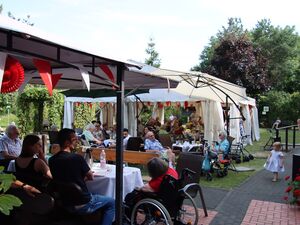 Geschmückte Festzelte, Draußen, Sonne scheint. Viele Menschen sitzen beisammen. Alte Menschen im Rollstuhl, alte Mensche mit Rollator in der Nähe, ein kleinem Mädchen im weißen Kleidchen kommt einen Weg auf die Kamera zu gelaufen.