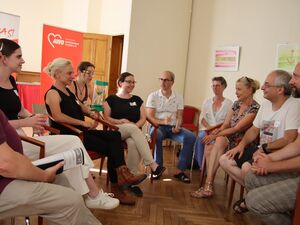 10 Menschen sitzen in einem Raum auf Stühlen. Eine Frau hat eine grüne sanduhr auf dem Schoß, die andere sitzen sich gegenüber.