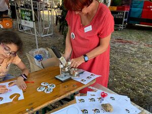 Buttons werden gemacht. Es gibt vorgedruckte kreisrunde Bilder, aber auch blanko Kreise zum selberbemalen. Mädchen sitzt am Tisch und malt. Frau mit roten Haaren und roten Kleid bedient die Button-Maschine.