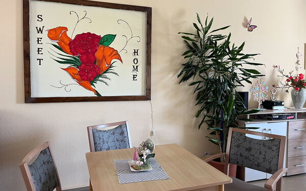 Gemeinschaftsraum: großes Bild mit Raosen und dem Text: SWEET HOME an der Wand. Davor ein Tisch um den drei Stühle stehen. Eine große Zimmerpflanze und ein Sideboard stehen an der Wand.