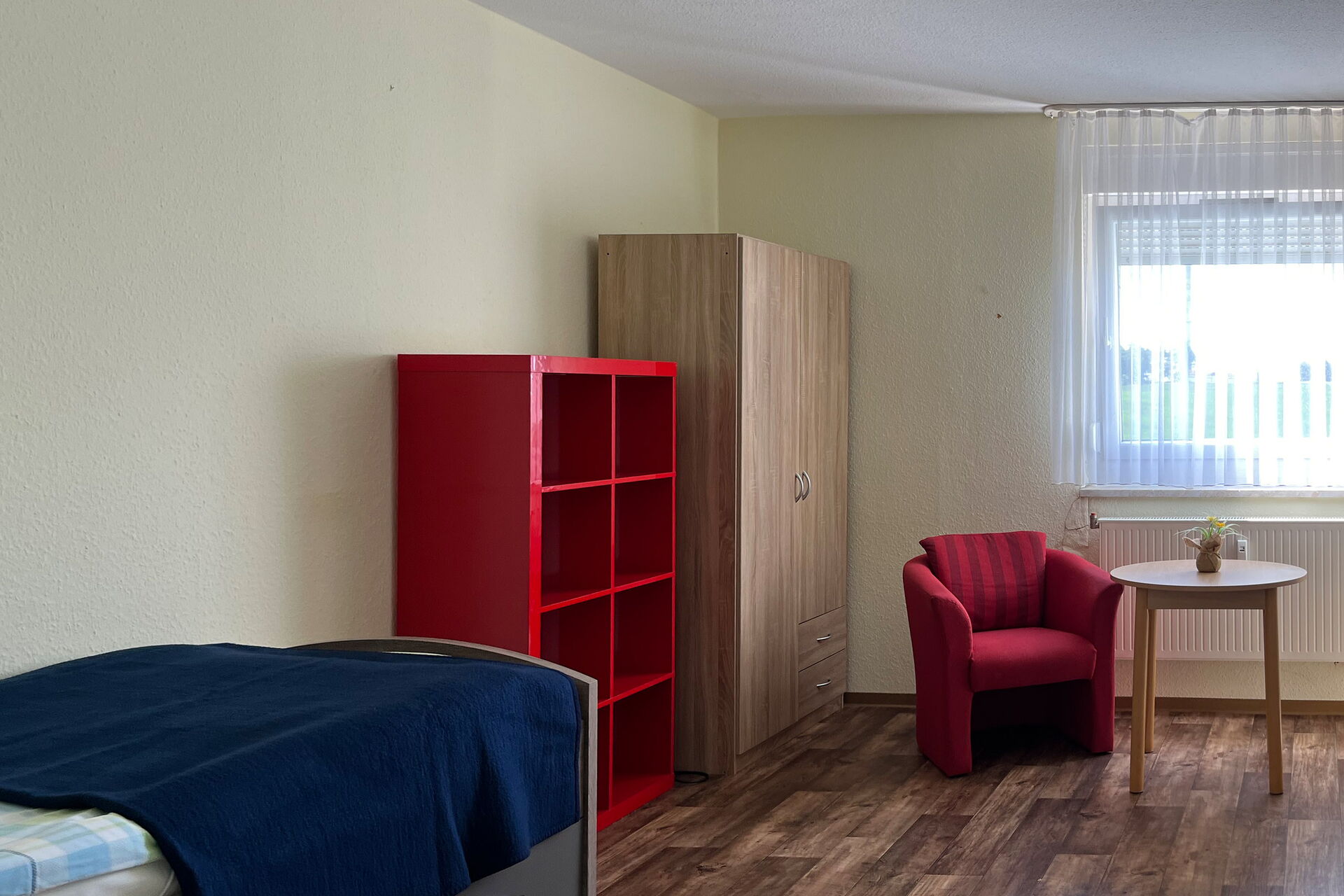 Bewohnerzimmer: vorn links ein Bett an der Wand weiter hinten ein rotes offenes Regal und ein Kleiderschrank in der Ecke des Zimmers. Ein roter Sessel und ein kleiner runder Tische stehen vor dem Fenster.
