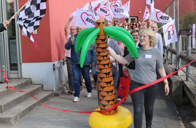 Eine Zielflagge links im Bild. Eine blonde Frau mit einer großen Gummi-Palme tritt über die Ziellinie (rotes Band) Hinter hier Teilnehmende im weißen AWO Flaggen.