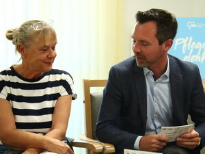 Frau mit blonden hochgesteckten Haaren und gestreiftem T-Shirt sitzt neben Mann in Anzug und Hemd, sie unterhalten sich.