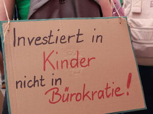 Frau mit grünem T-Shirt von hinten. Sie hat ein Schild umgehangen. Darauf steht: "Investiert in Kinder, nicht in Bürokratie!"