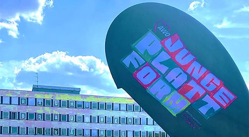 Gebäude, Rechenzentrum. Davor ein großer Sportplatz mit Basketballkorb und Toren. Die Sonne scheint. Wenige Personen sind auf dem Sportplatz. Werbefahne mit Aufdruck: AWO JUNGE PLATTFORM im Vordergrund.