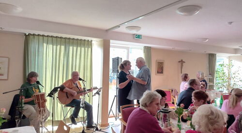 Heller Raum mit großen Fenstern. Kaffeetafeln und zwei Menschen die am Fenster sitzen und Musik machen. Rechts von dem Musikern ein tanzendes Paar.