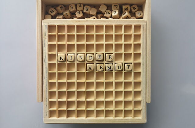 solide Holzbox mit Schiebedeckel beinhaltet Buchstabenwürfel, die in Wörter und Kreuzwortgebilde gelegt werden. Das Wort Kinderarmut ist über zwei Reihen gelegt.