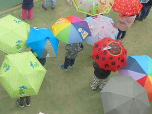 Personen, darunter Kinder an einem Sportplatz. Alle haben bunte Regenschirme über sich aufgespannt. Von oben fotografiert.