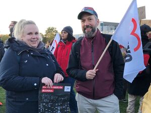 Füllige blonde Frau in schwarzer Winterjacke und Mann mit Basecap weinroter Jacke mit schwarzen Ärmeln. Der Mann schwängt eine weiße AWO Fahne. Im Hintergrund weitere demonstrierende Menschen..