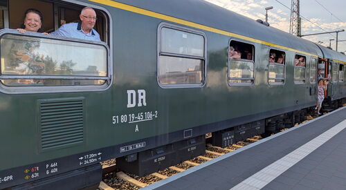 Dunkelgrüner Zug am Gleis, Menschen schauen aus den Fenstern.