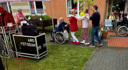 Polonäse: Draußen Sonne scheint. Vier Personen bilden eine Polonäse vor eine ältere Person im Rollstuhl.