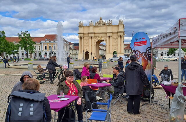 Luisenplatz Potsdam, das Brandenburger Tor im Hintergrund. Vorne Tische und Menschen die Kuchen essen und sich unterhalten. Ein Springbrunnen in der Mitte.