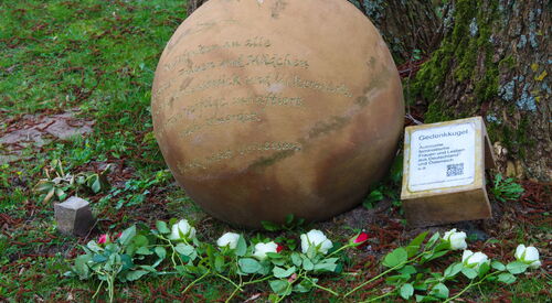 Draußen an einem Baumstamm ist eine Gedenkkugel. Davor liegen Blumen. Die Kugel besteht aus rot gebranntem Ton mit einer Inschrift:. Die Kugel hat eine kupferne Färbung.