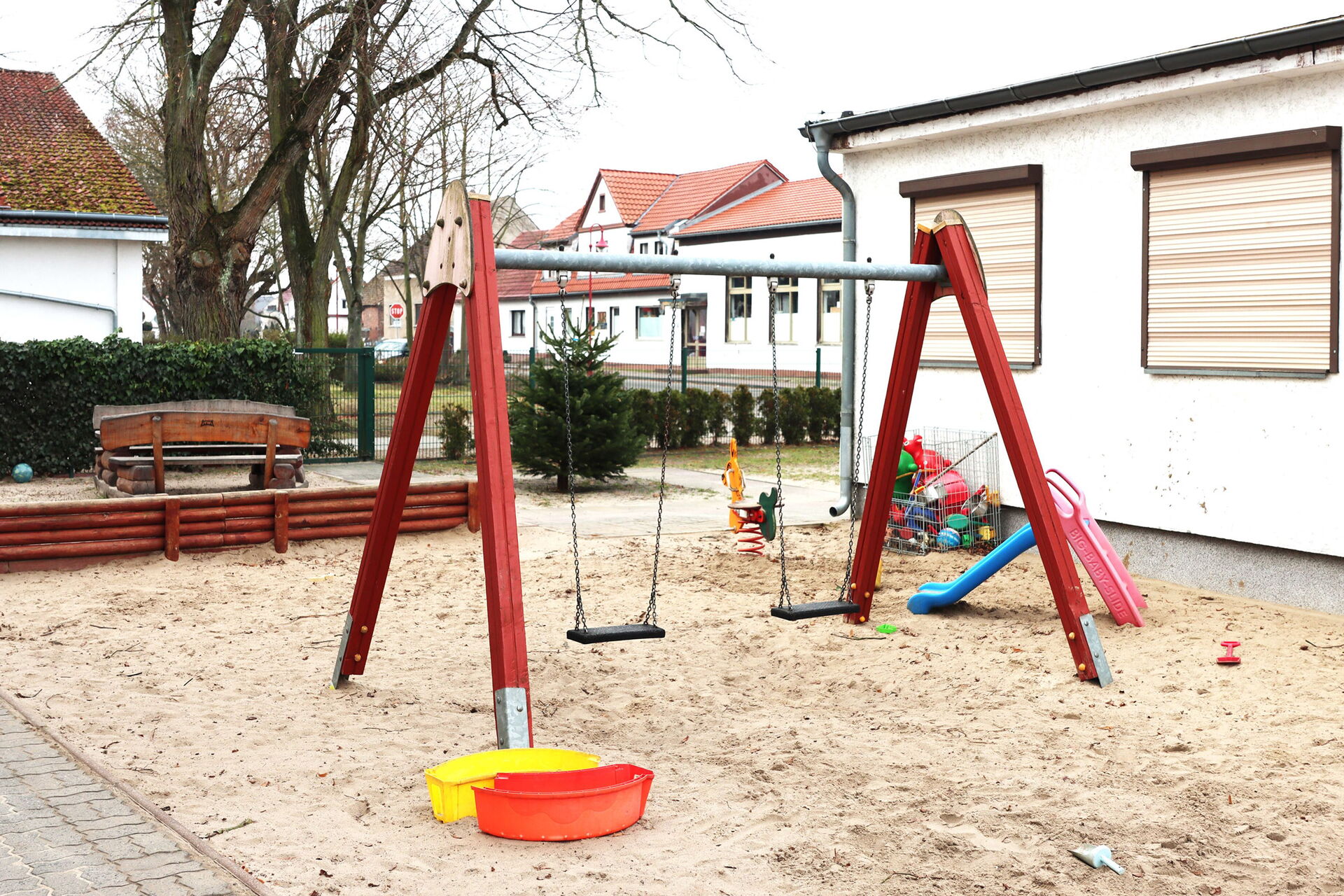 Großer Sandkasten mit zwei Schaukeln, Rutsche für Kleinkinder. Dahinter Holzbänke und eine Hecke.