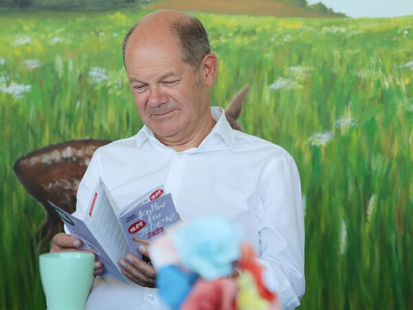 Mann sitzt vor einem Wandbild, dass eine weite Wiese mit Blumen zeigt. Mann guckt sich den Flyer von Kultur für Jeden an. Der Mann ist älter, hat eine Halbglatze, Haare an den Seiten sind kurz und grau. Er trägt ein weißes Hemd vor ihm eine Tasse.