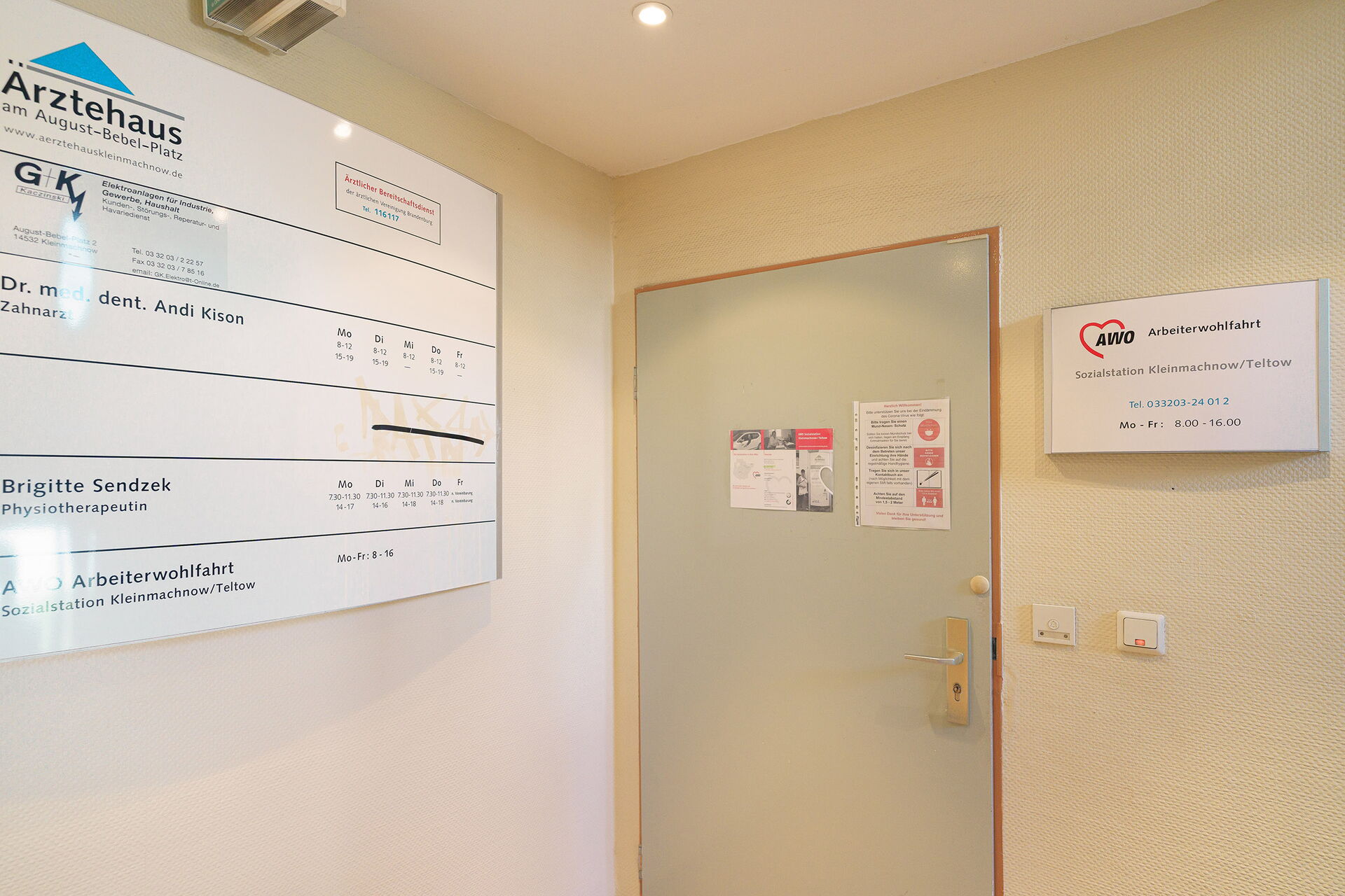 Flur. Geschlossene Tür zur Sozialstation. Links an der Wand ein großes Schild vom Ärztehaus worin sich die Sozialstation befindet.