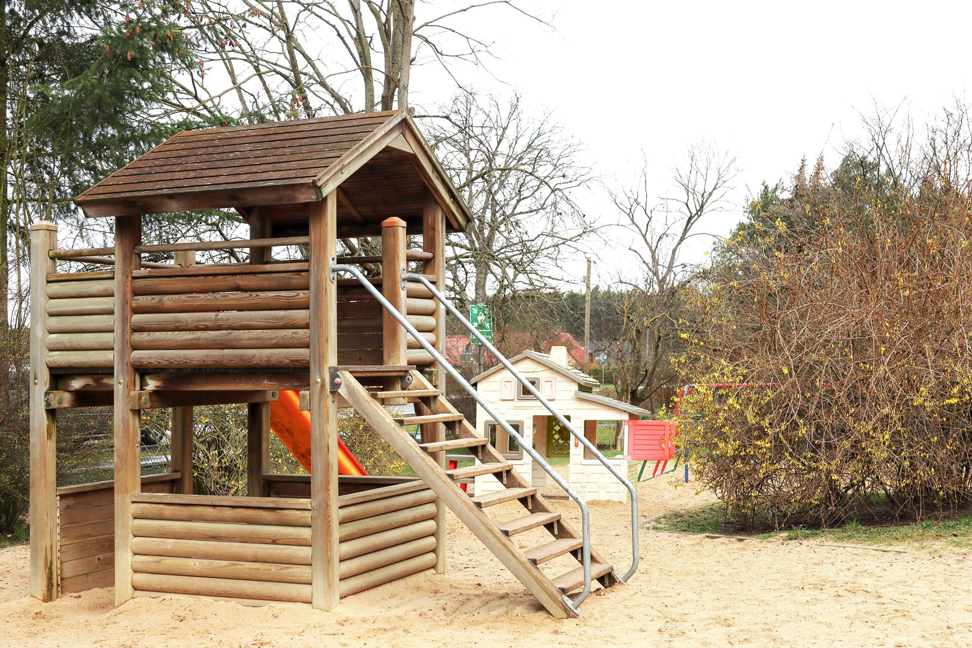 Holzklettergerüst mit Treppe und roter Rutsche im Sand.