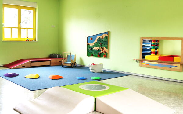 Spielzimmer. Großer Raum mit grünen Wänden. Auf einem blauen Teppich der in der Ecke liegt sind farbige Balanciersteine Für Kinder (lila, gelb, orange, blau, grün) in unterschiedlichen Größen An der Wand ein Spiegel.