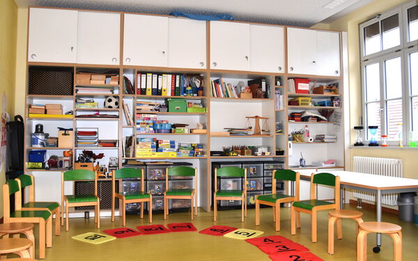 Ein Raum mit großen Regalen und Schränken, in den Regalen Brettspiele, Aktenordner.           Stühle und Hocker stehen in einem Halbkreis davor liegen große Plättchen mit Zahlen drauf.