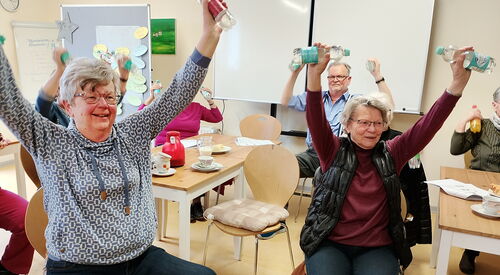 Zwei Seniorinnen sitzen auf Stühlen. Sie haben in jeder Hand eine kleine Flasche Wasser und heben die Arme nach oben. Sie lächeln. Hinter ihnen sitzen vier weitere Senior*inne und machen dieselbe Übung.