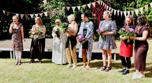 Gruppenbild: Team ganz links Frau mit Mikrofon. 7 Frauen halten Blumensträuße in den Händen.