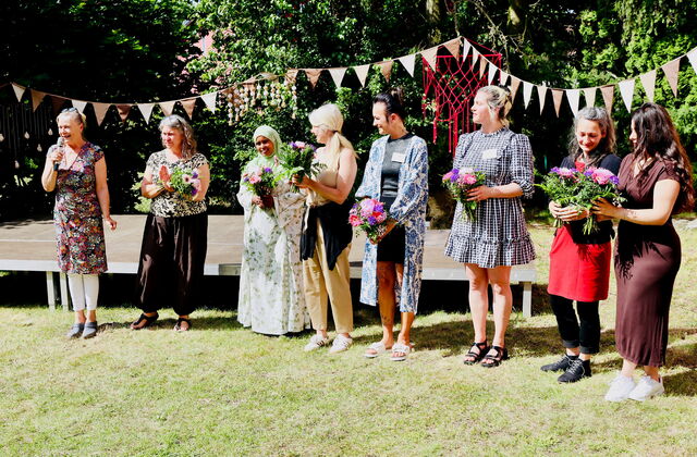 Gruppenbild: Team ganz links Frau mit Mikrofon. 7 Frauen halten Blumensträuße in den Händen.