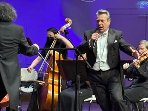 Orchester mit Dirigent und einem Tenor auf der Bühne. Alle schwarz gekleidet. Tenor in Anzug und weißem Hemd und weißer Fliege.