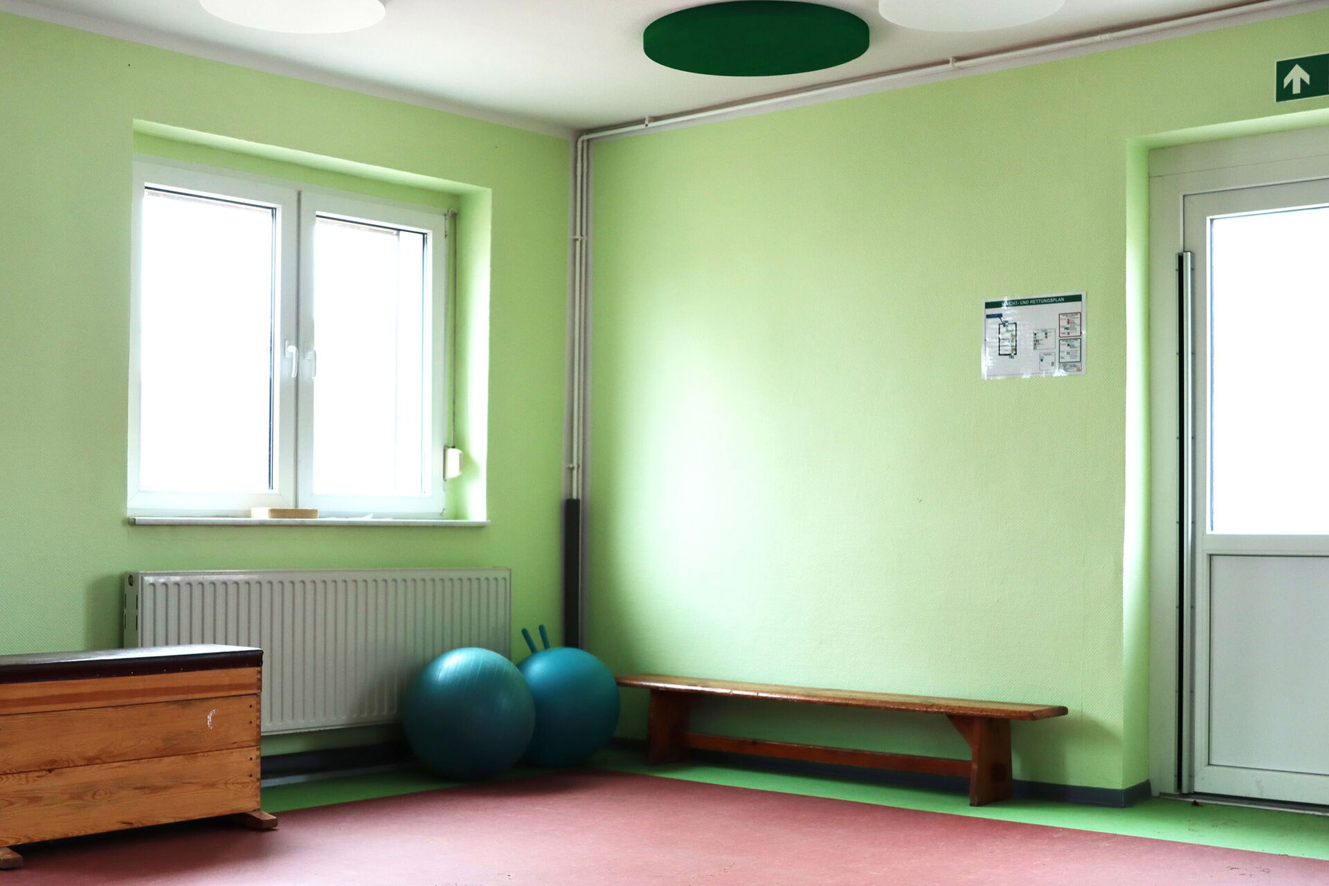 Turnhalle. Grüne Wände. Ein Fenster, eine weiße Tür, mit Notausgang-Schild darüber. Eine Bank steht an der Wand, zwei Pezzi-Bälle und ein Sprungkasten