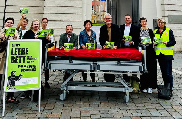 vor dem Fortuna Portal. Ein leeres Pflegebett, ein großes grünes Schild und 10 Menschen mit grünen Karten, mit Aufschrift.