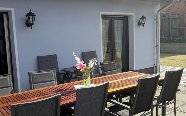 Große Terrasse mit langen Tisch und Stühlen vor einem Haus. Auf dem Tisch steht eine Vase mit Blumen. Die Sonne scheint.