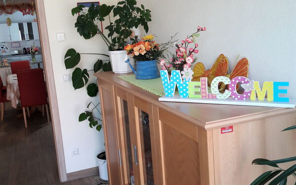 Flur. Eine Kommode an der Wand rechts. Darauf Blumen und ein buntes Schild: WELCOME.