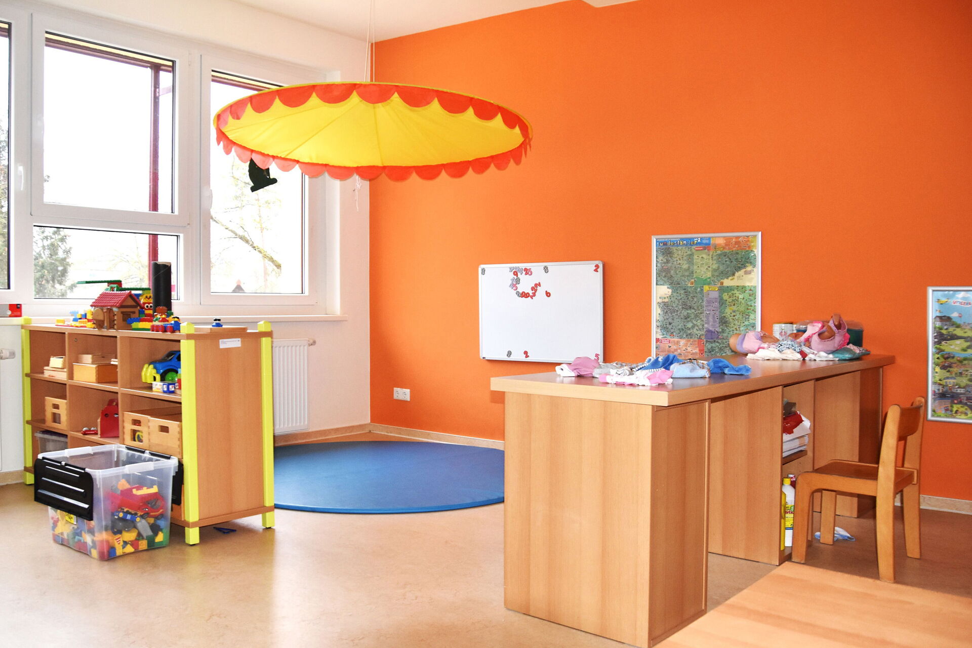 Spielzimmer. Freundlich eingerichteter Raum für Kinder. Die Wand ist orange, Regal mit Lego und Spielzeug links am Fenster.