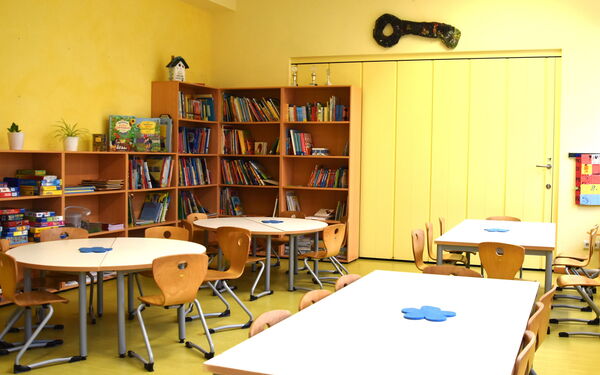Runde und rechteckige Tische mit Stühlen und große Regale in der Ecke und an der linken Wand. In den Regalen Brettspiele und Bücher.