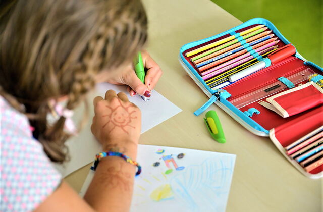 Mädchen sitzt am Tisch und schreibt mit der linken Hand. Auf dem Tisch liegen eine Federmappe zum aufklappen mit Buntstiften, Linial und Anspitzer. Und ein gemaltes Bild liegt unter dem rechten Arm des Mädchens.
