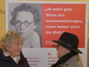 Hintergrund: Plakat mit schwarz-weiß Foto von Marie Juchacz mit Zitat: Je mehr gute Taten wir zusammenbringen, umso besser wird die Welt. Davor zwei ältere Frauen im Gespräch.